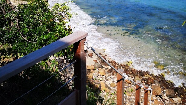 Une vue côtière avec une plage dégagée, un escalier en bois descendant vers le rivage et, en arrière-plan, des collines parsemées de bâtiments.