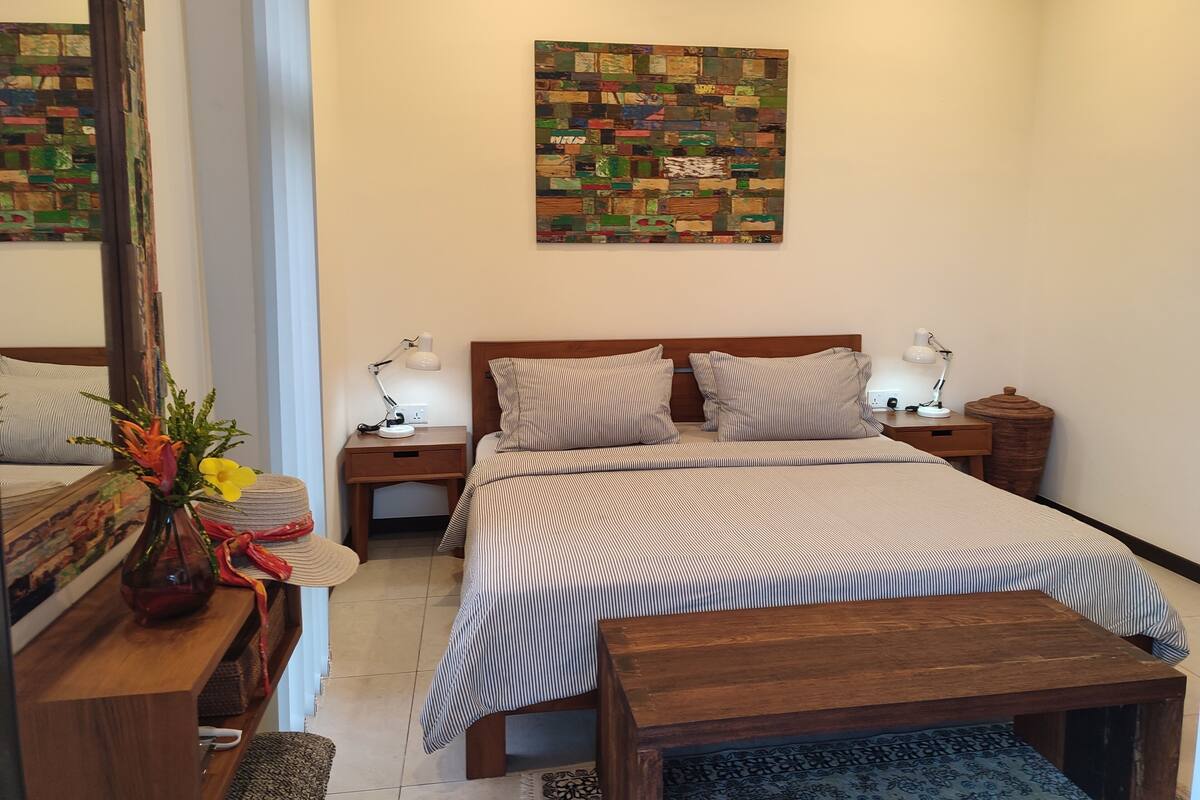 Quarto de hotel com uma cama, duas mesas de cabeceira com luminárias, um banco de madeira, um vaso decorativo com flores e uma obra de arte colorida na parede.
