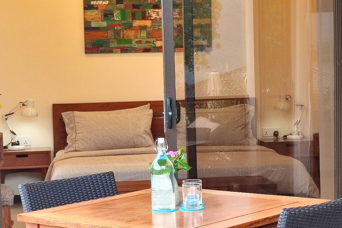 Quarto de hotel com uma cama, uma mesa de madeira com uma garrafa e copos, e um quadro na parede.