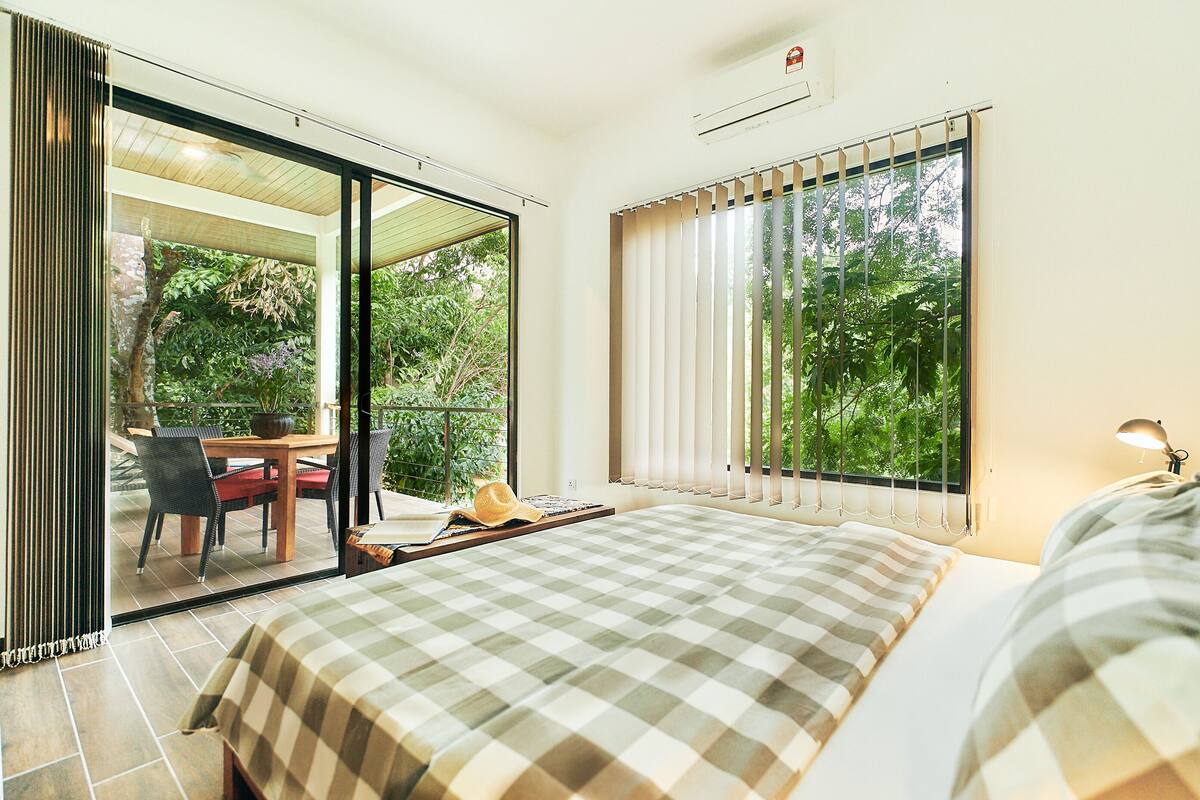 Quarto com colcha xadrez, escrivaninha com cadeira e vista para o verde pela janela.