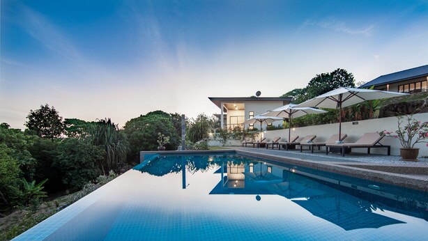 A modern pool area with lounge chairs and umbrellas, surrounded by lush greenery and a building with a sloped roof. - Ranis Lodge (Langkawi)