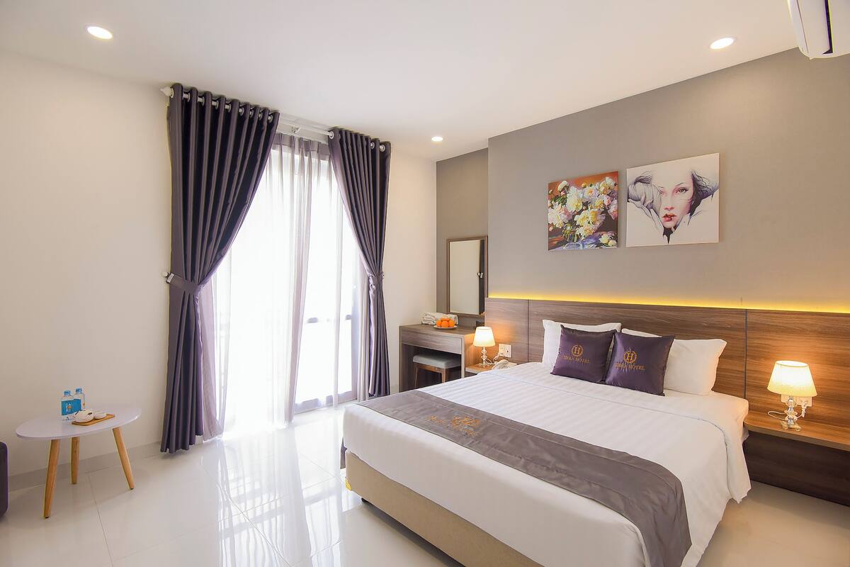A modern hotel room with a large bed, bedside tables with lamps, a small desk, and a view through the curtains.