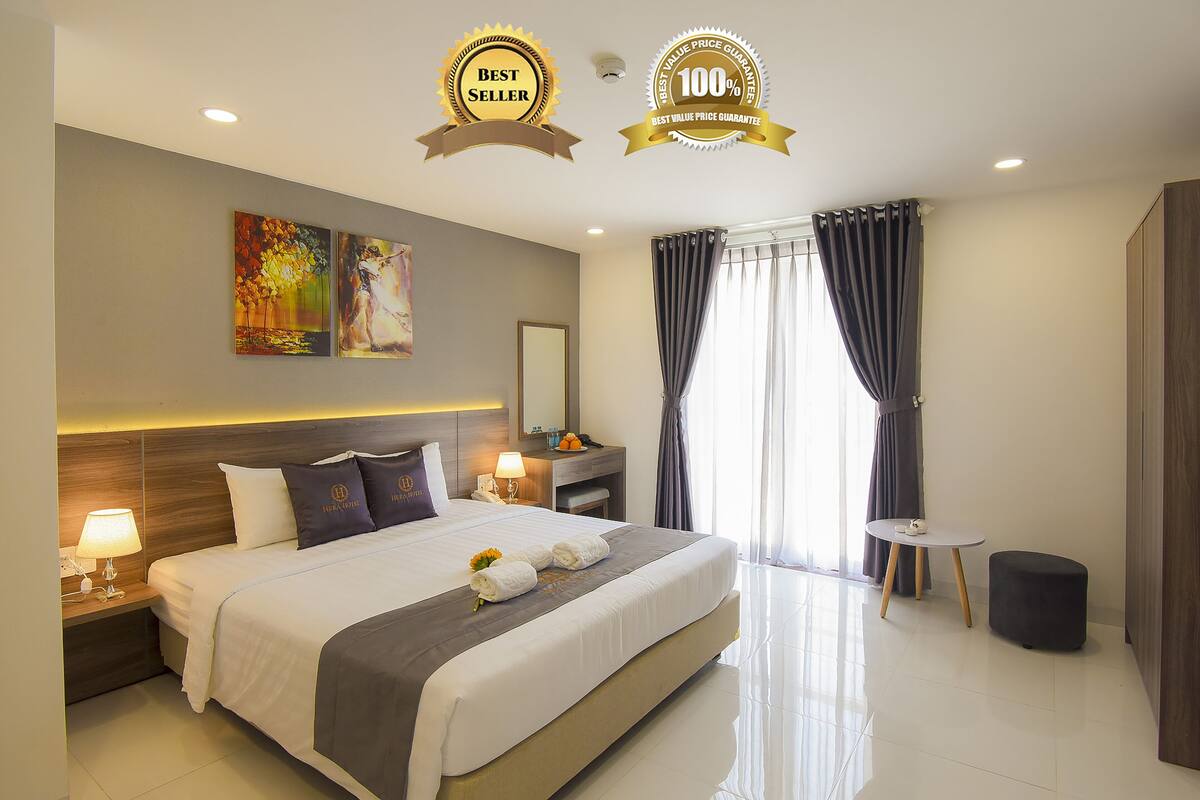 A modern hotel room with a large bed, bedside tables with lamps, a small round table, and a large window with curtains.
