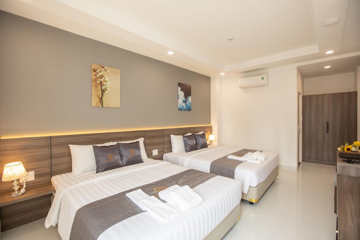 A hotel room with two beds, a wooden headboard, a wall-mounted air conditioner, and a small table with flowers.