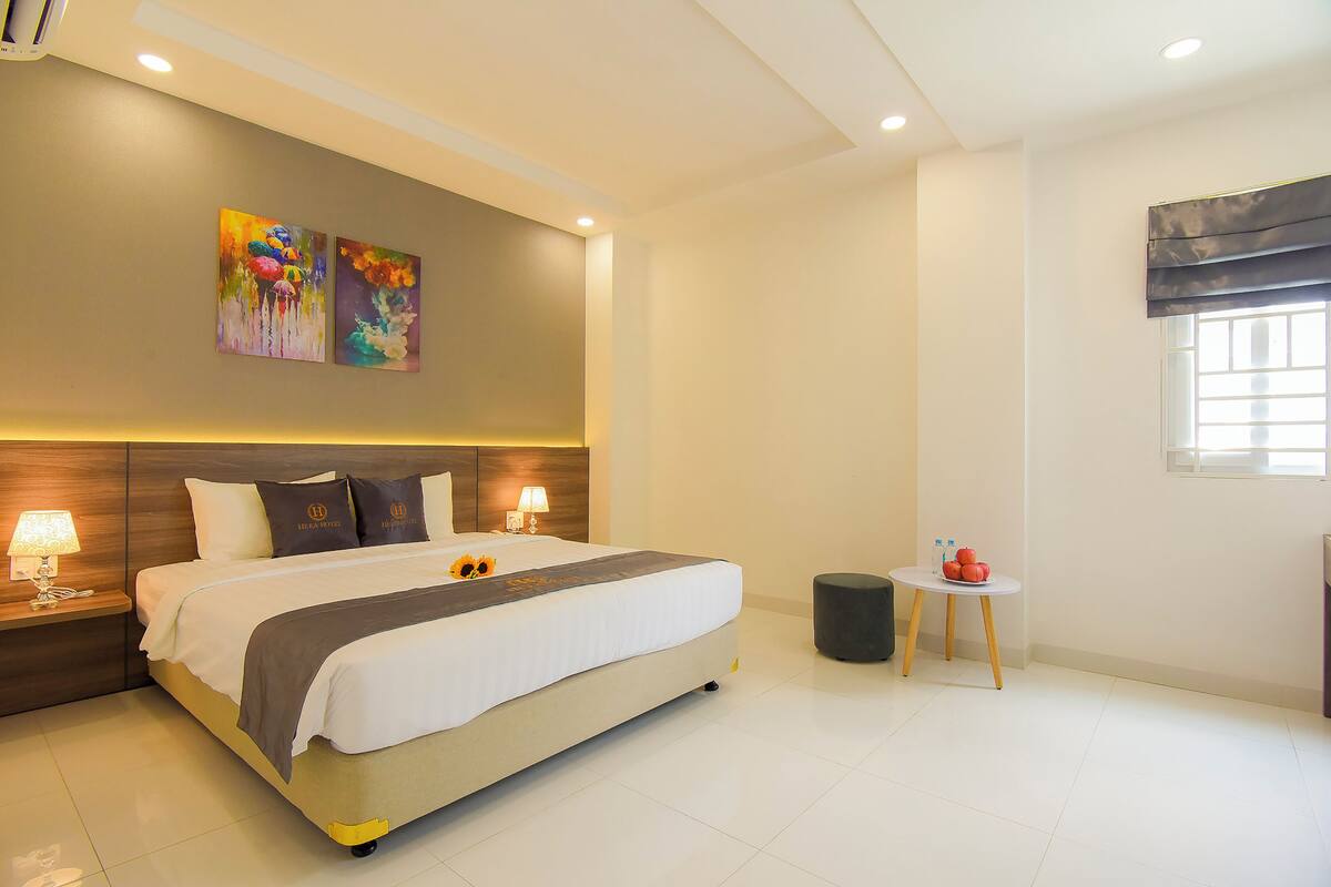 A modern hotel room with a large bed, bedside lamps, a small table with a fruit bowl, and two paintings on the wall.