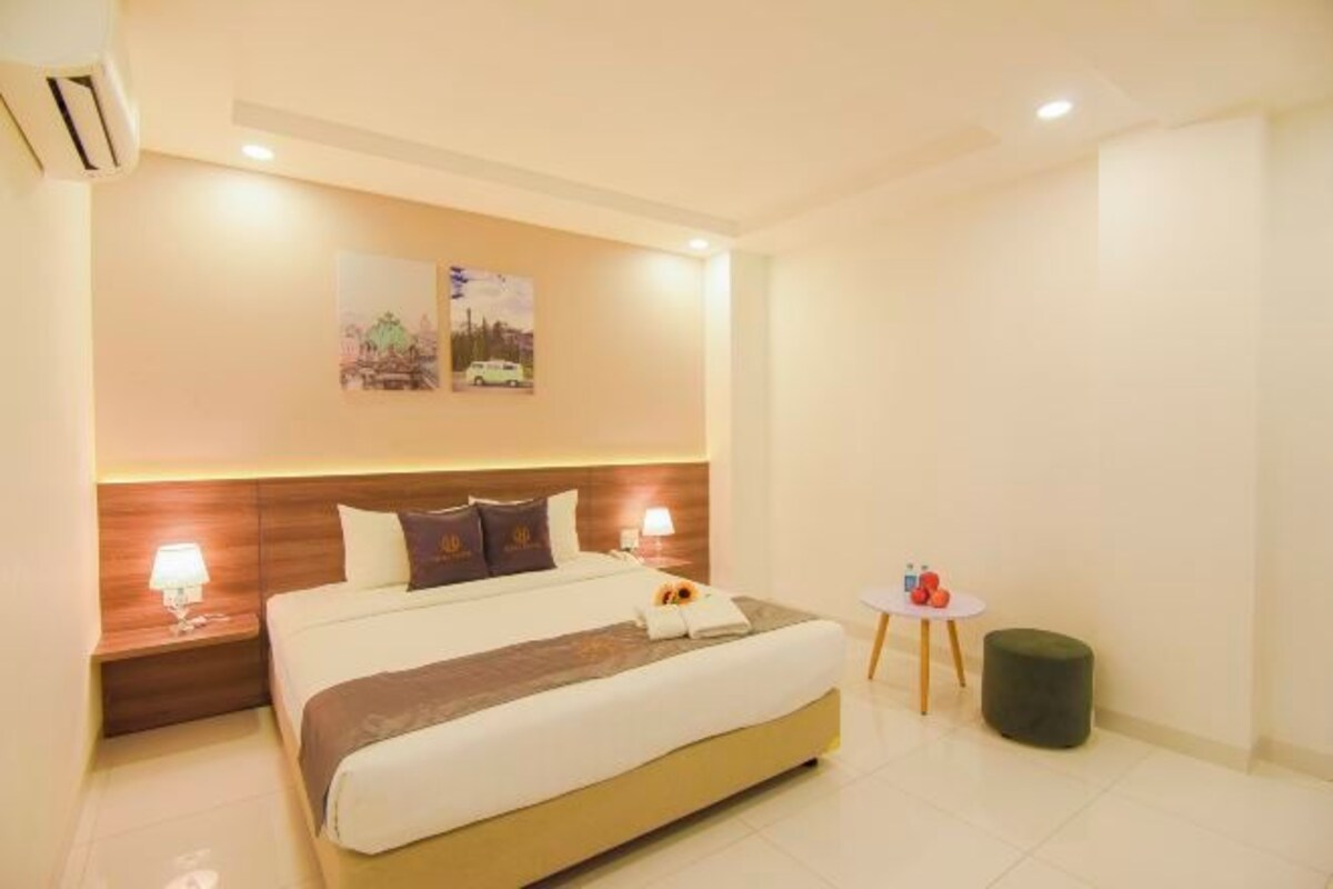 A hotel room with a large bed, two bedside tables with lamps, a small round table with a red object, and a green ottoman.