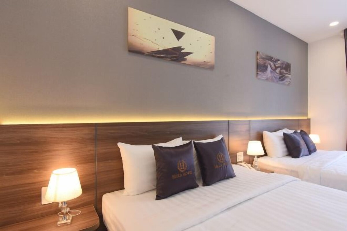 A hotel room with a wooden headboard, two beds with white linens, dark pillows with a logo, a wall-mounted artwork, and a nightstand with a lamp.