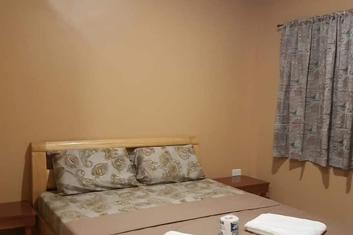 A single bed with a floral bedspread, two pillows, a bedside table, and a window with patterned curtains.