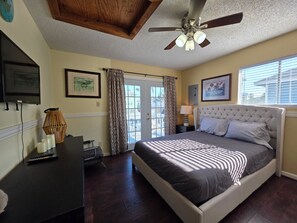 A bedroom with a bed, a ceiling fan, a desk with a lamp, and a TV.