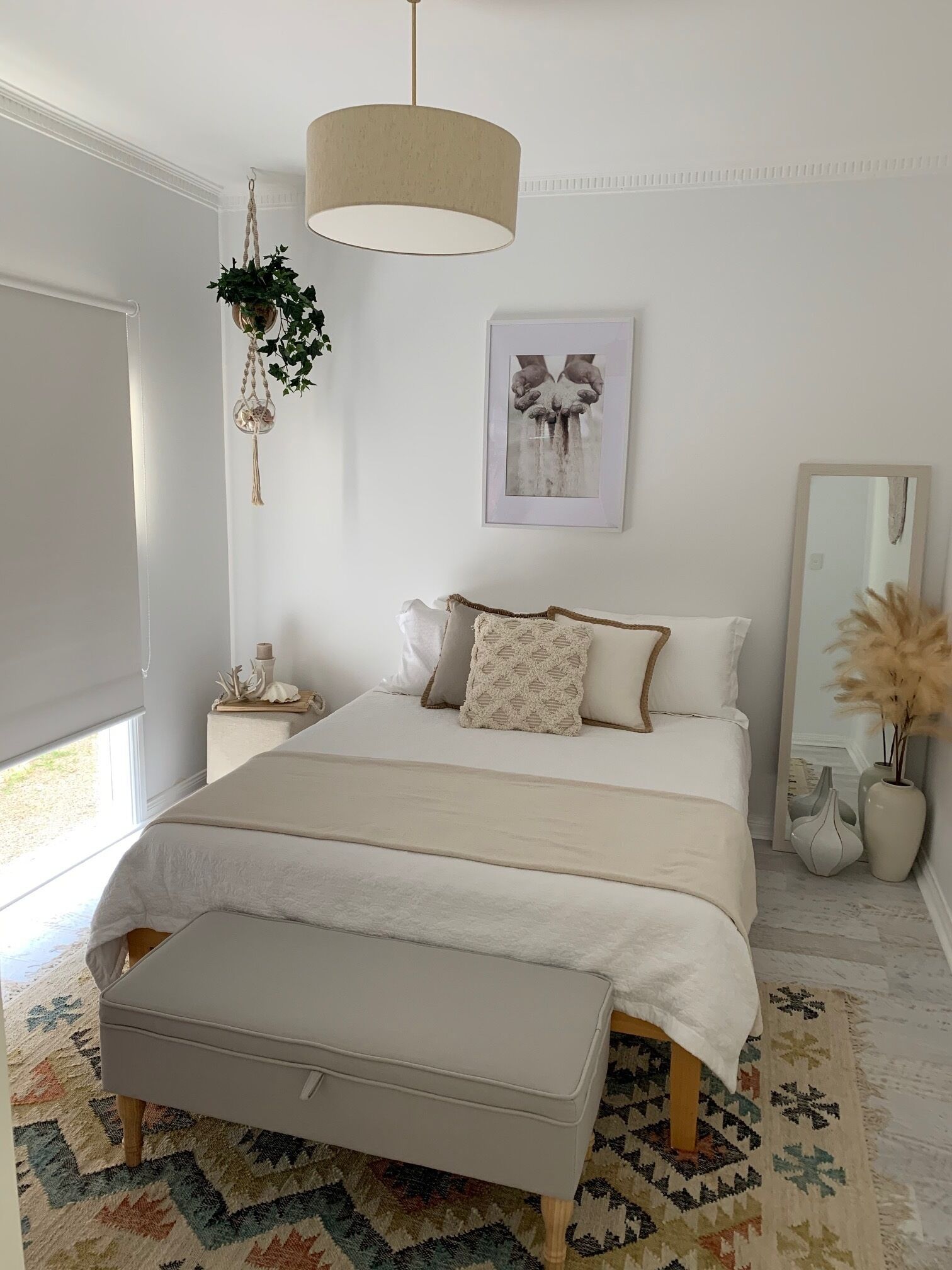 A bedroom with a bed, bedside tables, a hanging plant, a framed picture, a mirror, and a patterned rug.