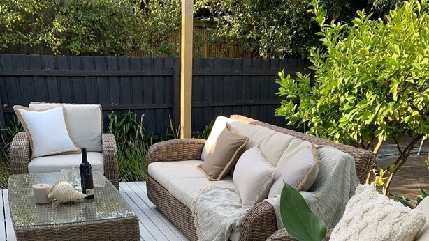 Un espace extérieur accueillant, aménagé avec des meubles en osier, dont un canapé et un pouf, entouré de plantes en pot et agrémenté d’une table en bois sur laquelle sont disposés une bougie et des objets décoratifs.