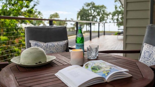 Een houten tafel met een boek, een kaars, een fles en een glas op een balkon met uitzicht op bomen en een balustrade.