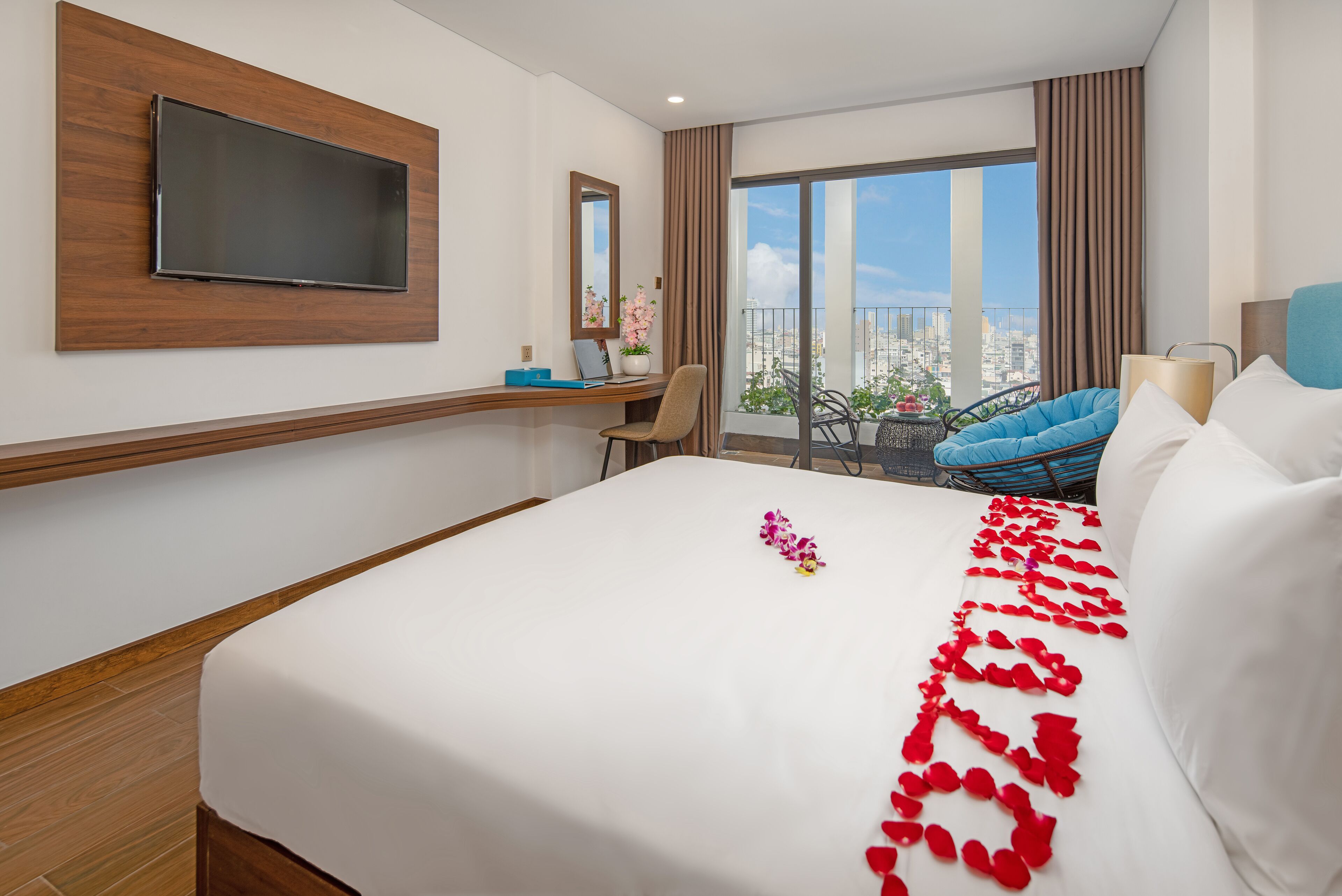 A hotel room with a large bed, a desk with a laptop, a TV mounted on the wall, a balcony with a chair, and a city view.