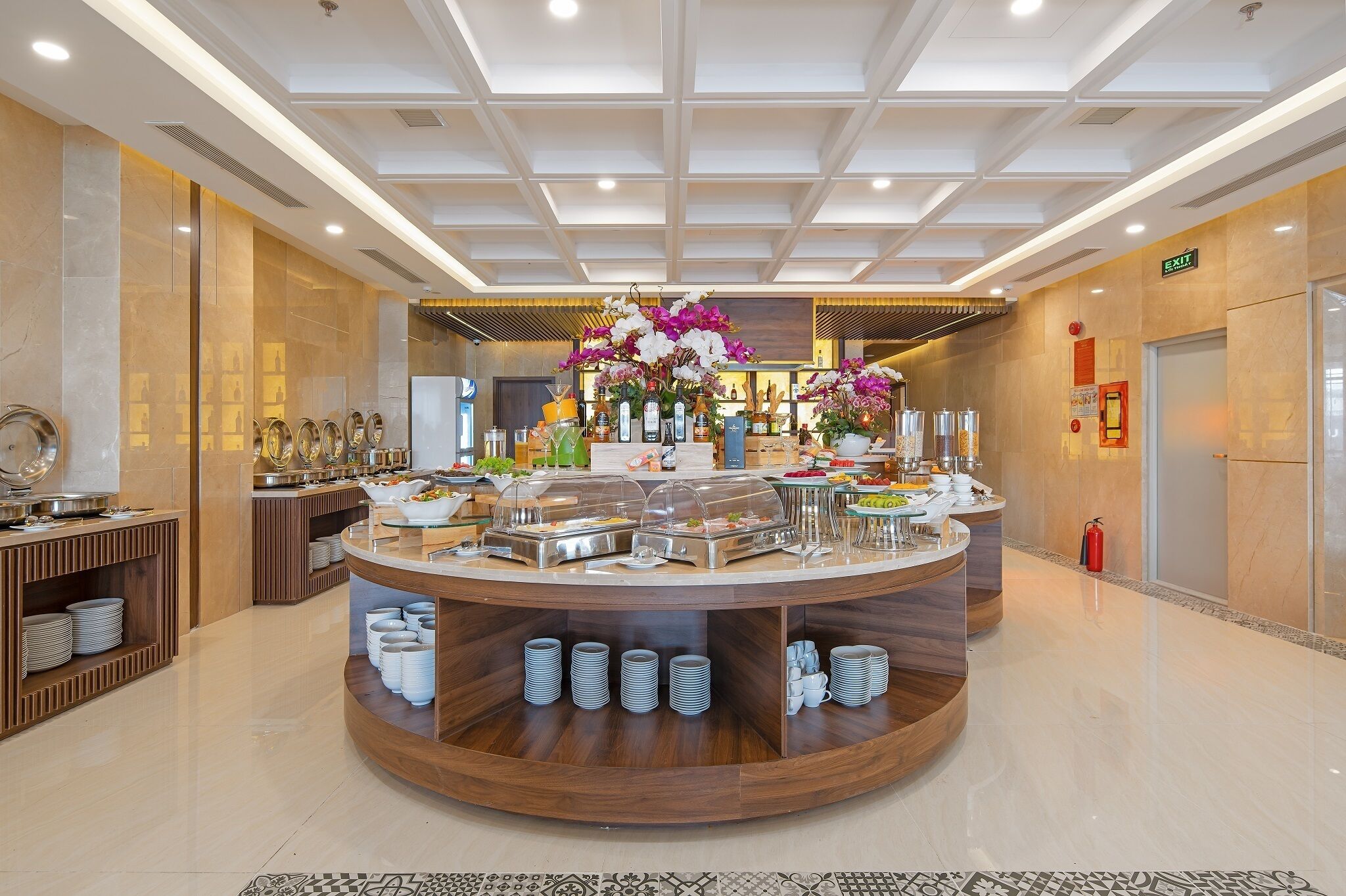 A modern hotel lobby with a central buffet area featuring food and drink dispensers, a decorative floral arrangement, and a polished marble floor.