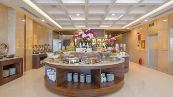 A modern hotel lobby with a central buffet area featuring food and drink dispensers, a decorative floral arrangement, and a polished marble floor.