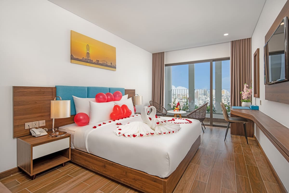 A modern hotel room with a large bed, a desk, and a balcony with a city view.