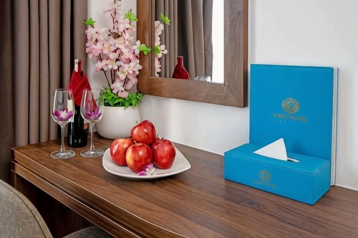 Una mesa de madera con un plato de manzanas, dos copas de vino, un florero con flores y una caja azul.