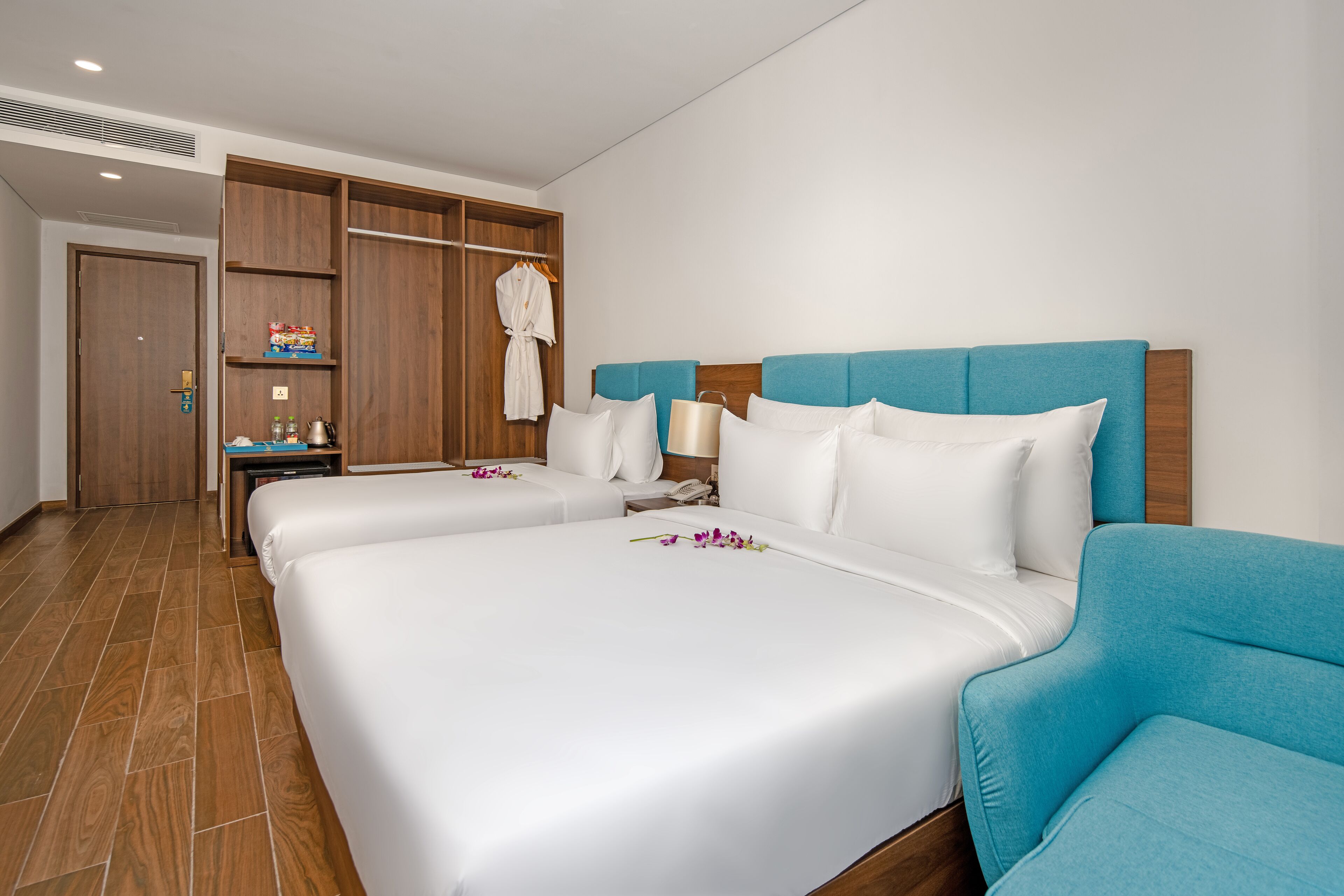 A hotel room with two beds, a wooden wardrobe, a blue armchair, and a nightstand with a lamp.