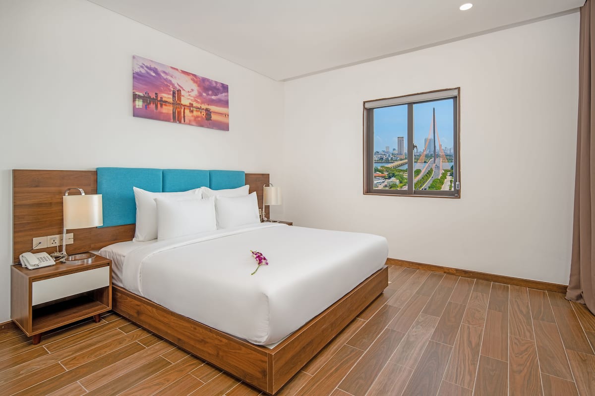 A hotel room with a large bed, a nightstand, a lamp, a painting, and a window with a cityscape view.