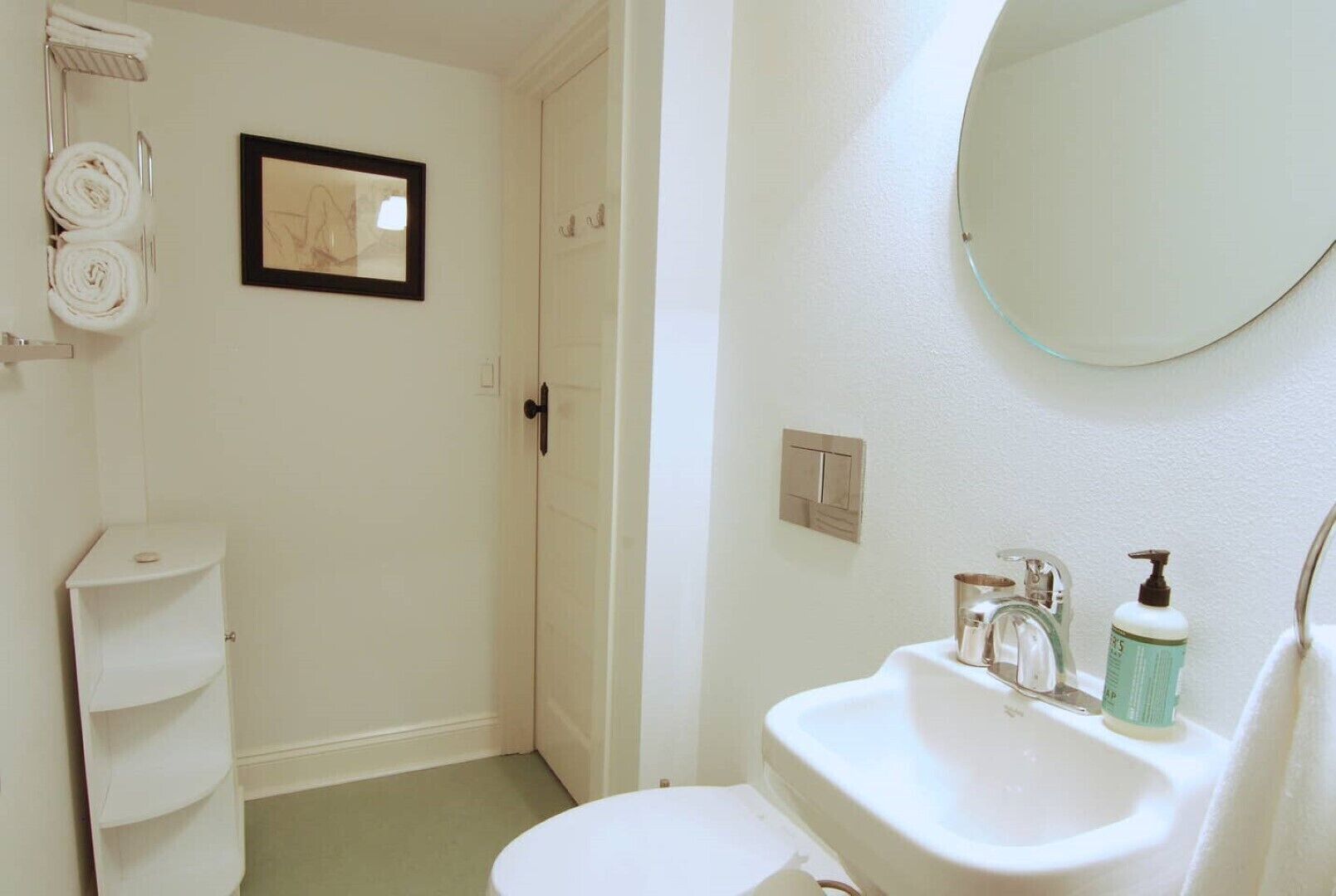 A bathroom with a toilet, sink, and mirror. There is a towel rack with towels and a framed picture on the wall.