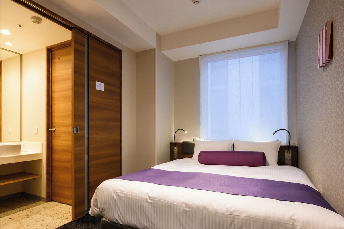 Een hotelkamer met een bed, een raam, een nachtkastje, een lamp en een badkamer met een wastafel en spiegel.