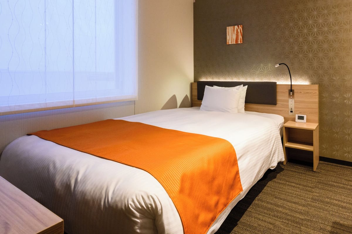 Een hotelkamer met een net opgemaakt bed, een nachtkastje met een lamp, een klein klokje en een raam met luxaflex.