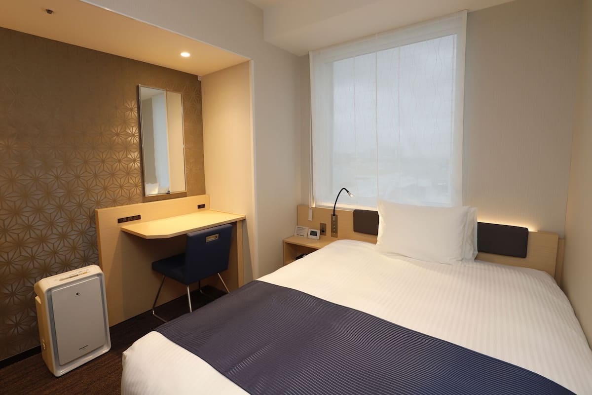 Een hotelkamer met een bed, een bureau, een stoel, een kleine koelkast, een nachtkastje, een lamp en een raam met dunne gordijnen.