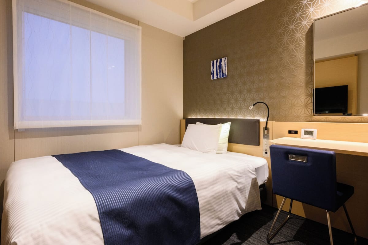 Een hotelkamer met een bed, bureau, stoel, raam met zonwering, wandklok en spiegel.