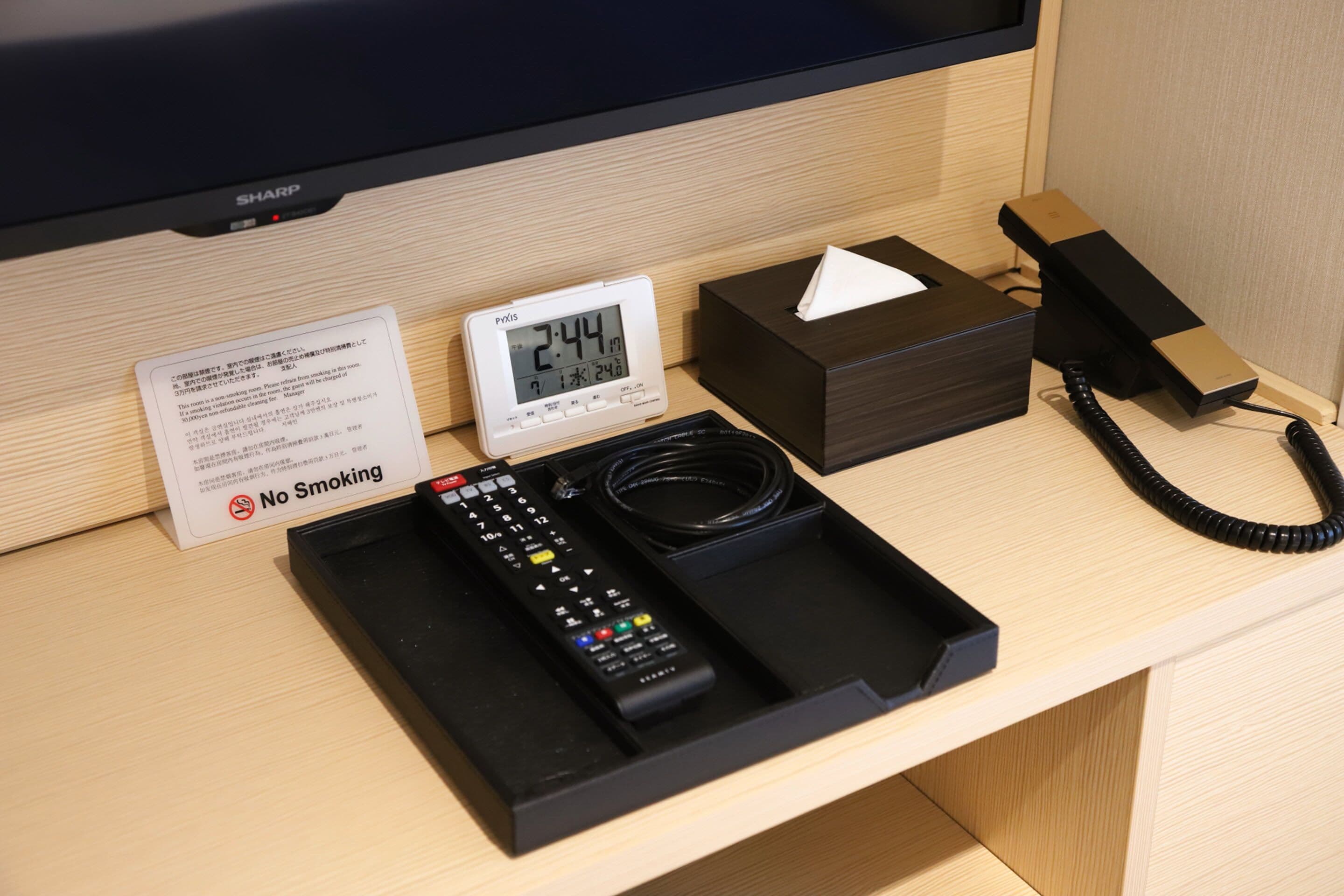 Habitación de hotel con una mesita de noche que contiene un reloj digital, una caja de pañuelos, un teléfono y un control remoto.