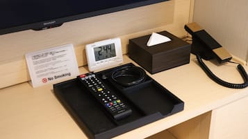 Habitación de hotel con una mesita de noche que contiene un reloj digital, una caja de pañuelos, un teléfono y un control remoto.