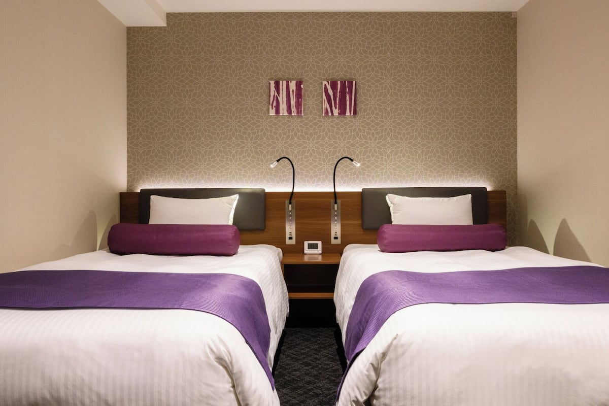 Een hotelkamer met twee bedden, elk met een paars deken en witte lakens. Daarboven hangt een kunstwerk aan de muur en daartussen staat een klein tafeltje.