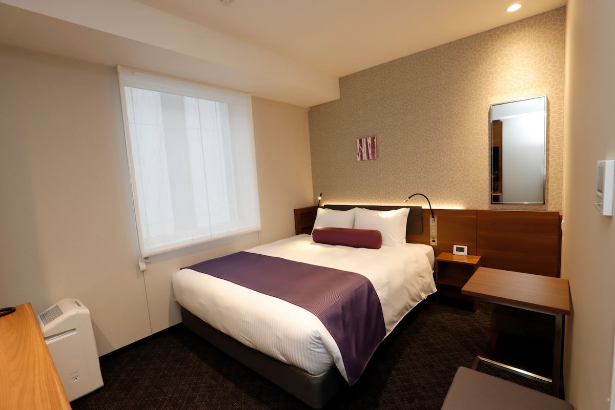 Een hotelkamer met een bed, nachtkastje, raam en een spiegel.