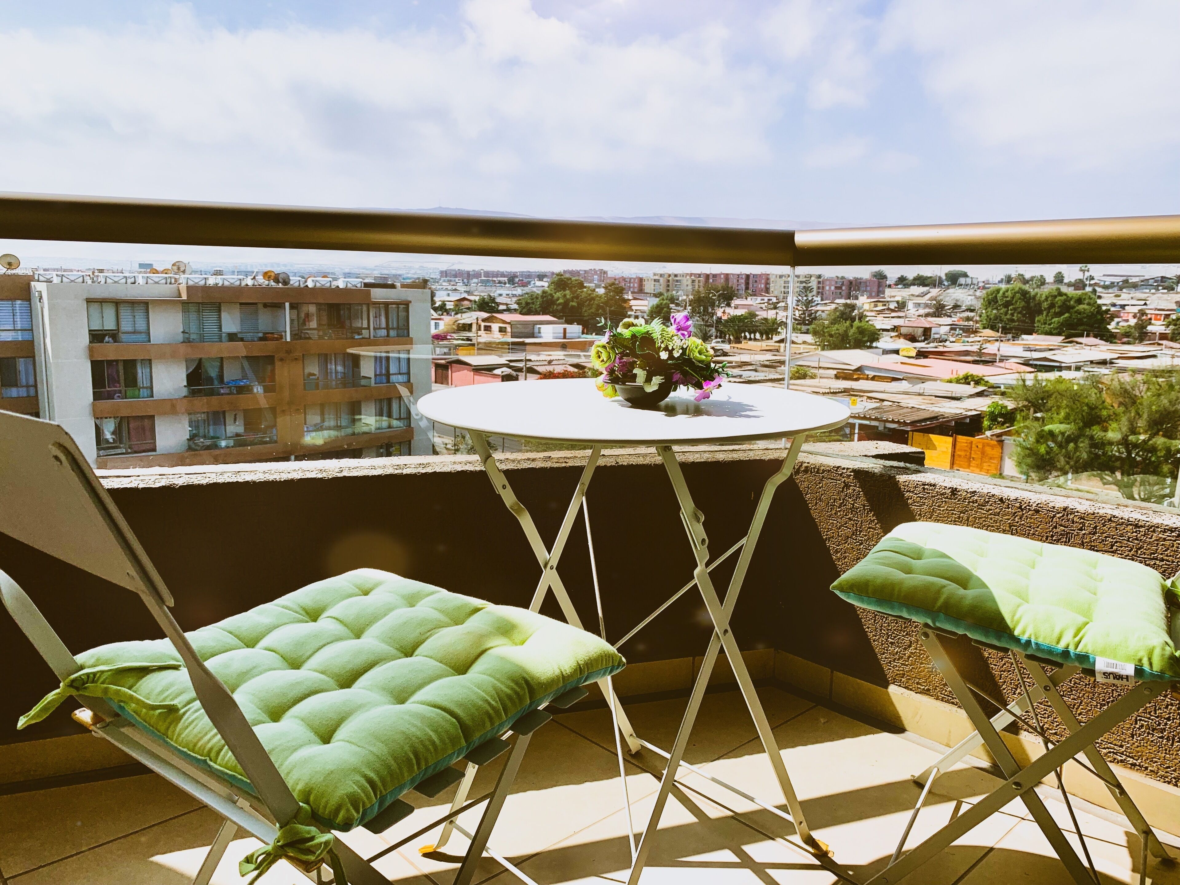 Ein Balkon mit zwei grünen Sitzgelegenheiten, einem kleinen runden Tisch mit Blumenvase und Blick auf eine Stadtansicht.