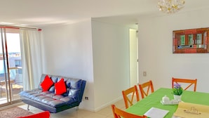Ein modernes Wohnzimmer mit einer Couch, Sesseln, einem Esstisch mit grüner Tischdecke und einem Kronleuchter.