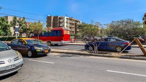 Straßenszene mit einem roten Bus, einem blauen Auto und einem silbernen Auto. Im Hintergrund sind Gebäude und Bäume zu sehen.