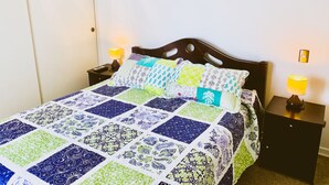 Ein ordentlich bezogenes Bett mit einer farbenfrohen Patchworkdecke, zwei Nachttischlampen und einem kleinen gerahmten Bild an der Wand.