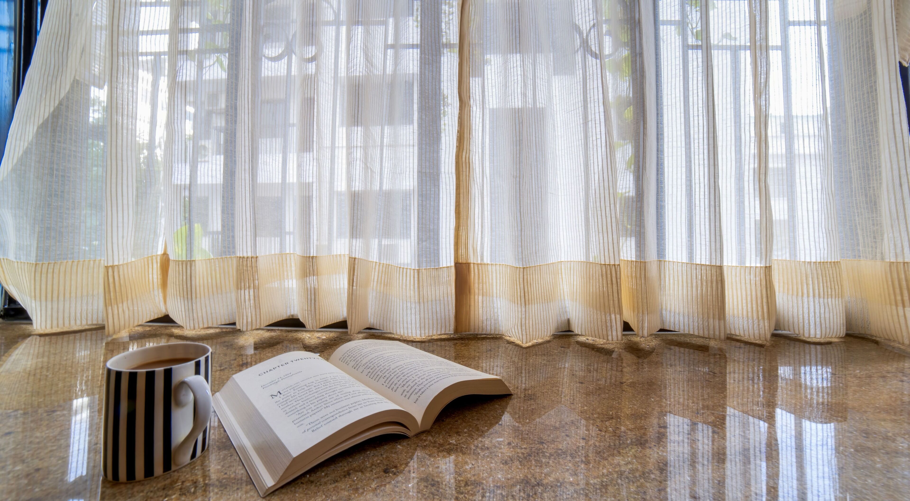 Ein Buch liegt offen auf einem Marmorboden, daneben eine gestreifte Tasse. In der Nähe ist ein Fenster mit durchsichtigen Vorhängen.