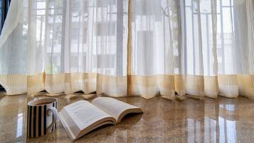 Ein Buch liegt offen auf einem Marmorboden, daneben eine gestreifte Tasse. In der Nähe ist ein Fenster mit durchsichtigen Vorhängen.