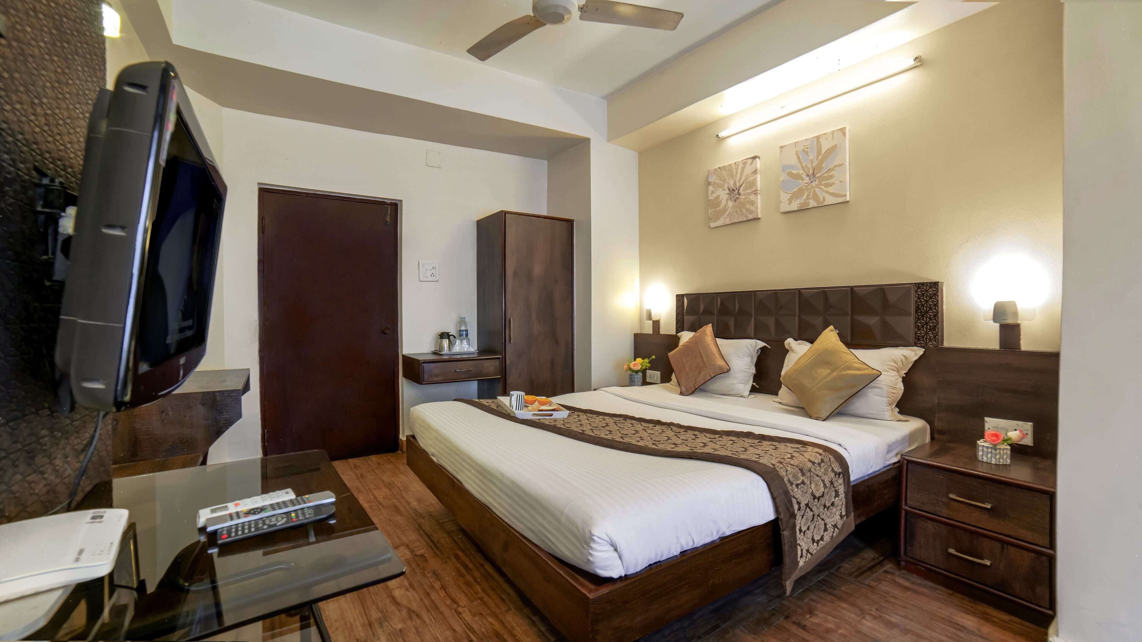 A hotel room with a bed, bedside tables, a TV, a desk with a computer and remote controls, a wardrobe, and artwork on the wall.