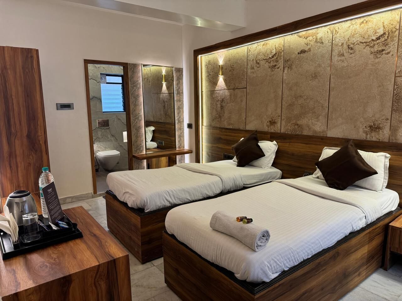 Habitación de hotel con dos camas, cabecero de madera, espejo y una mesita con un set de amenities.