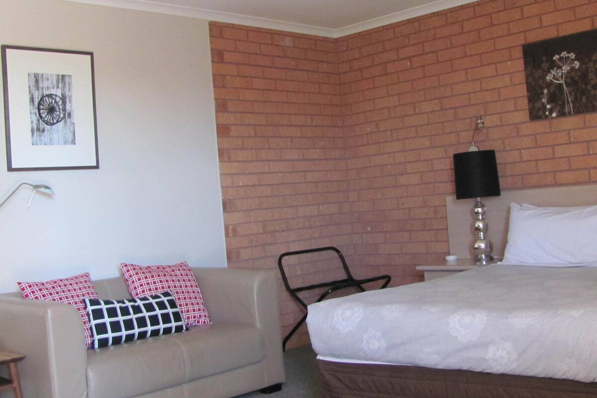 A hotel room with a brick wall, a bed with white linens, a beige sofa with patterned cushions, a side table, and a floor lamp.