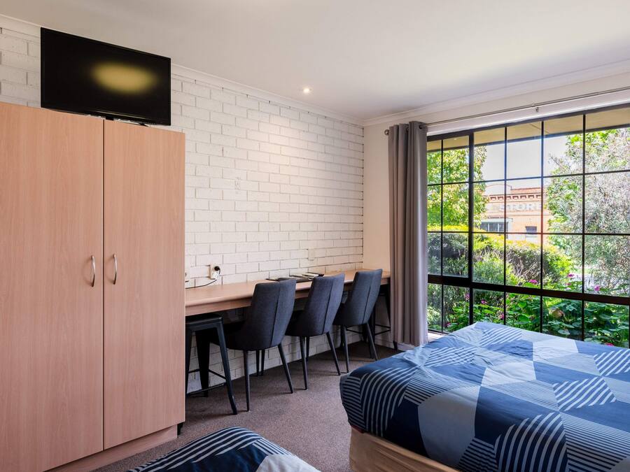A room with a bed, a desk with chairs, a TV mounted on a brick wall, and a window with a view of greenery.