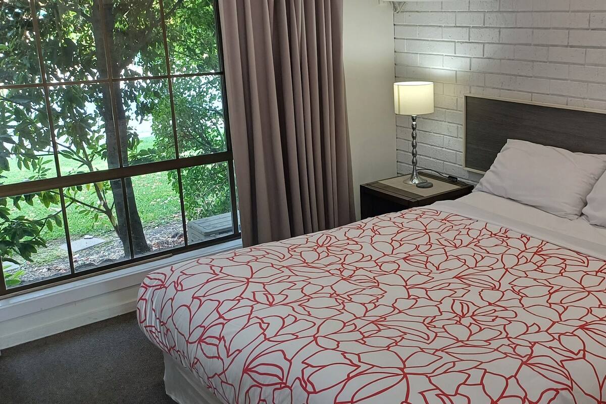 A hotel room with a large window, a bed with a patterned bedspread, a nightstand with a lamp, and a white brick wall.