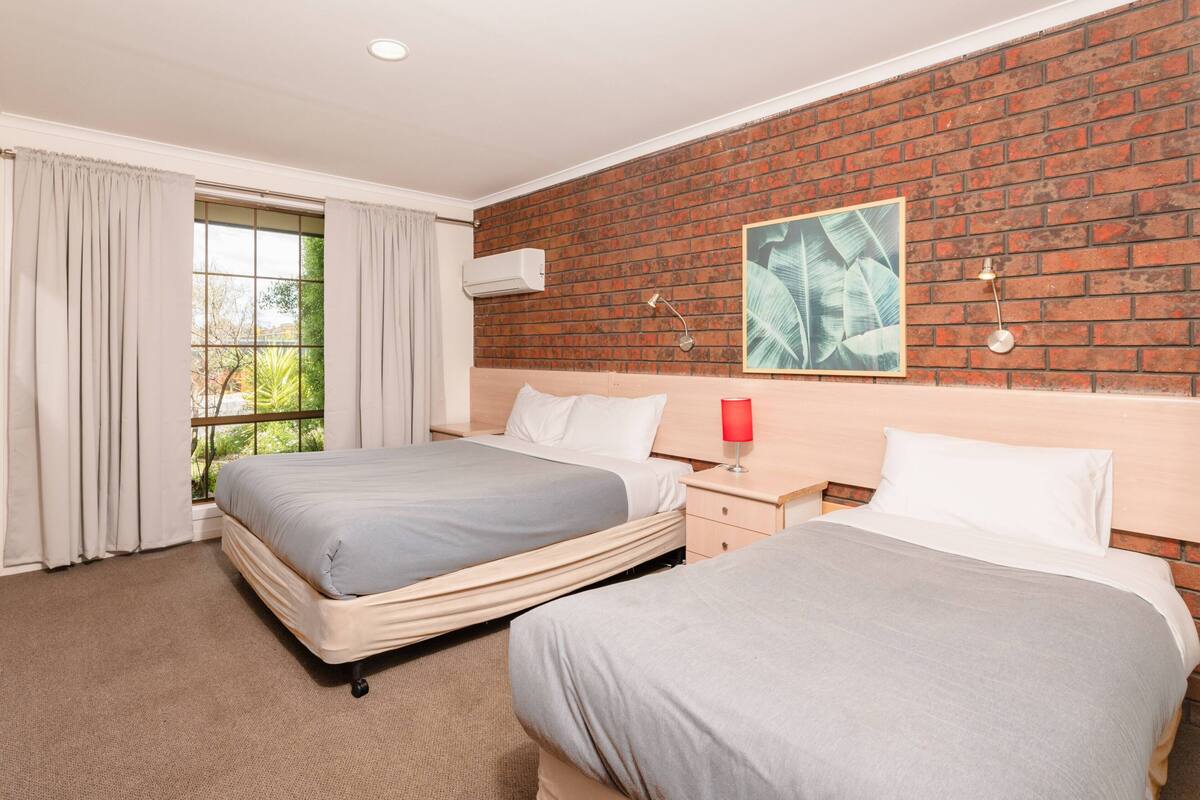 A hotel room with two beds, a brick wall, a window with curtains, a nightstand, and a wall-mounted lamp.