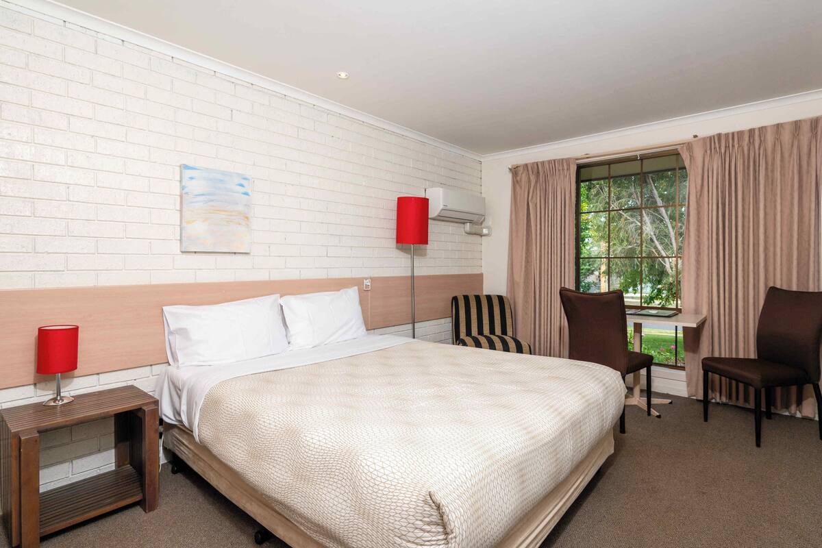A hotel room with a large bed, a desk, two chairs, a window with curtains, a red lamp, and a wall-mounted air conditioner.