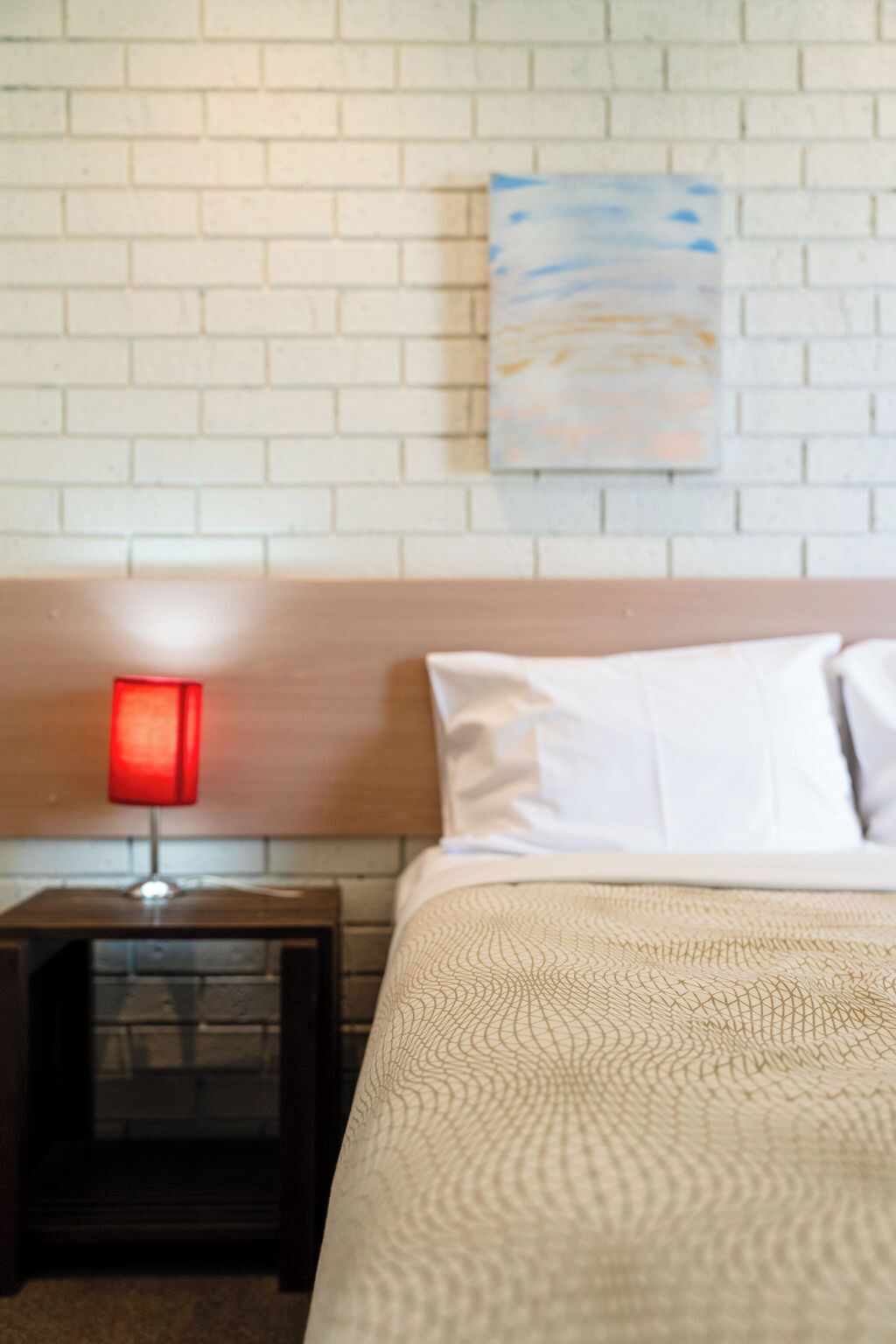 Quarto de hotel com cama, mesa de cabeceira com abajur vermelho, parede de tijolos brancos e um quadro.