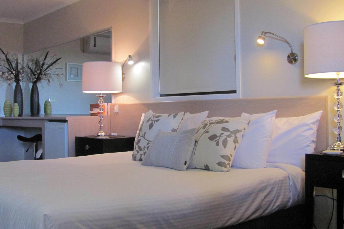 A hotel room with a bed, a nightstand, a lamp, a mirror, and a vase with flowers on the dresser.