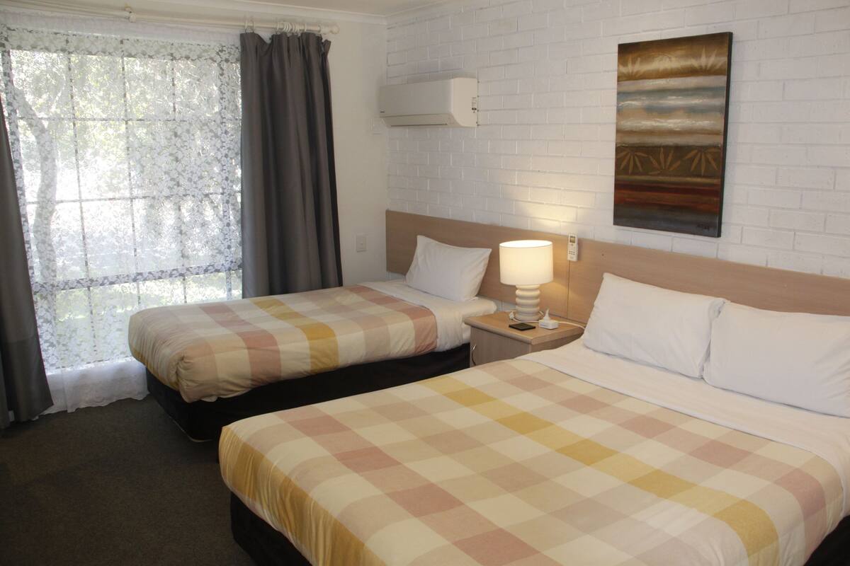 A hotel room with two beds, a window with curtains, a wall-mounted air conditioner, and a painting.