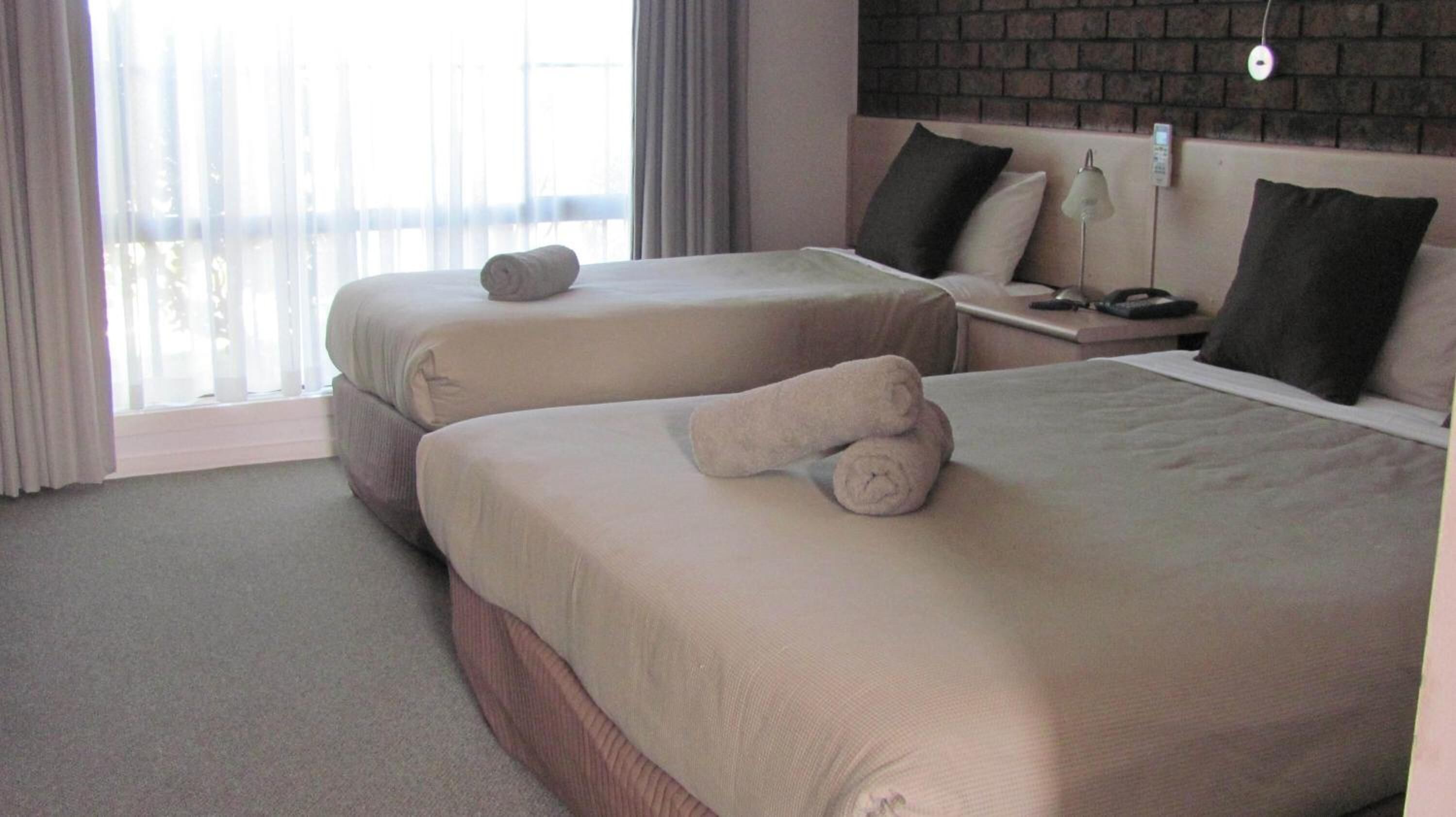 A hotel room with two beds, each with a rolled towel. There is a brick wall behind the beds, a nightstand with a lamp, and a window with curtains.