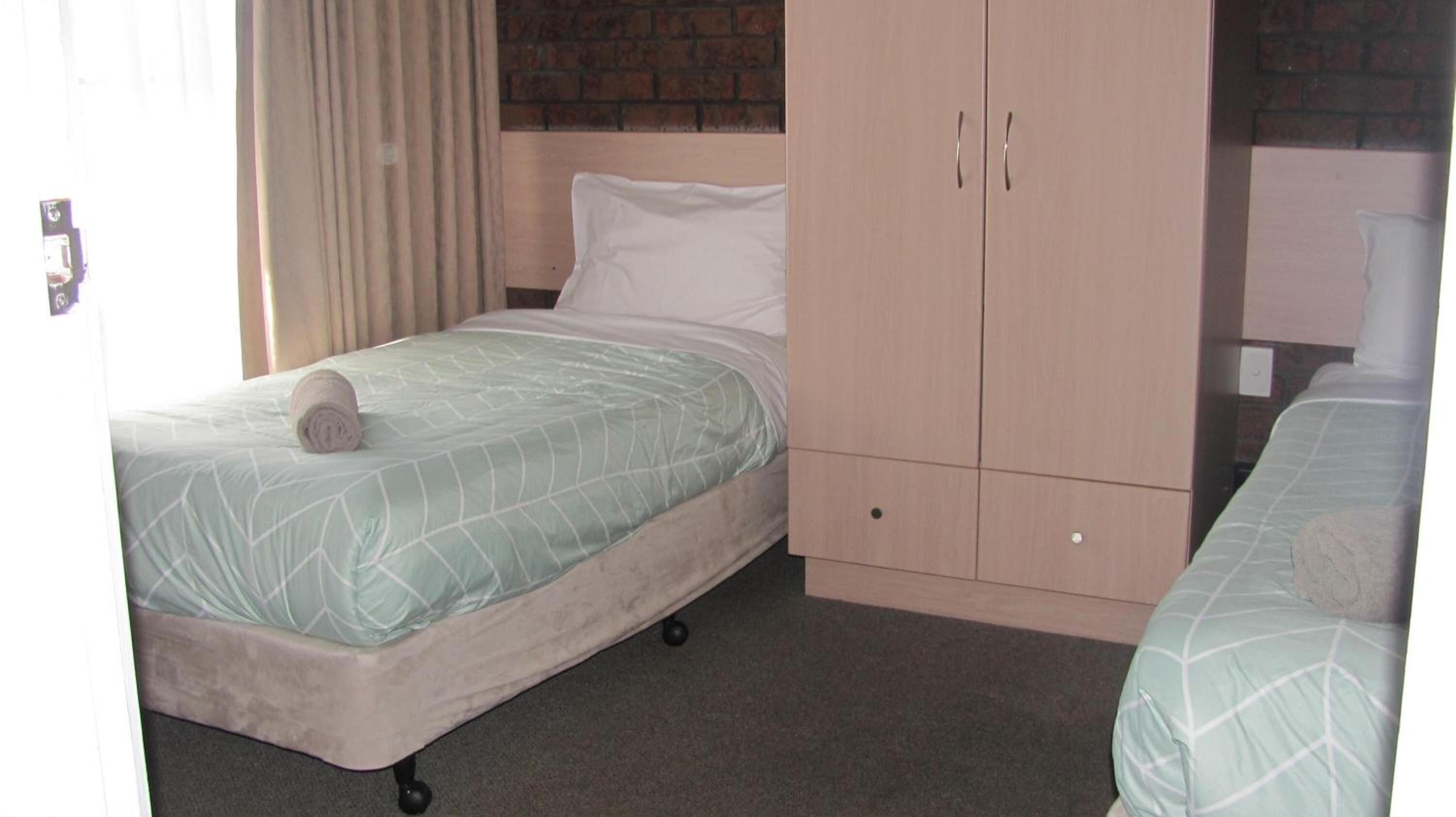 Een enkel bed met een opgerolde handdoek, een houten garderobe en een bakstenen muur op de achtergrond.